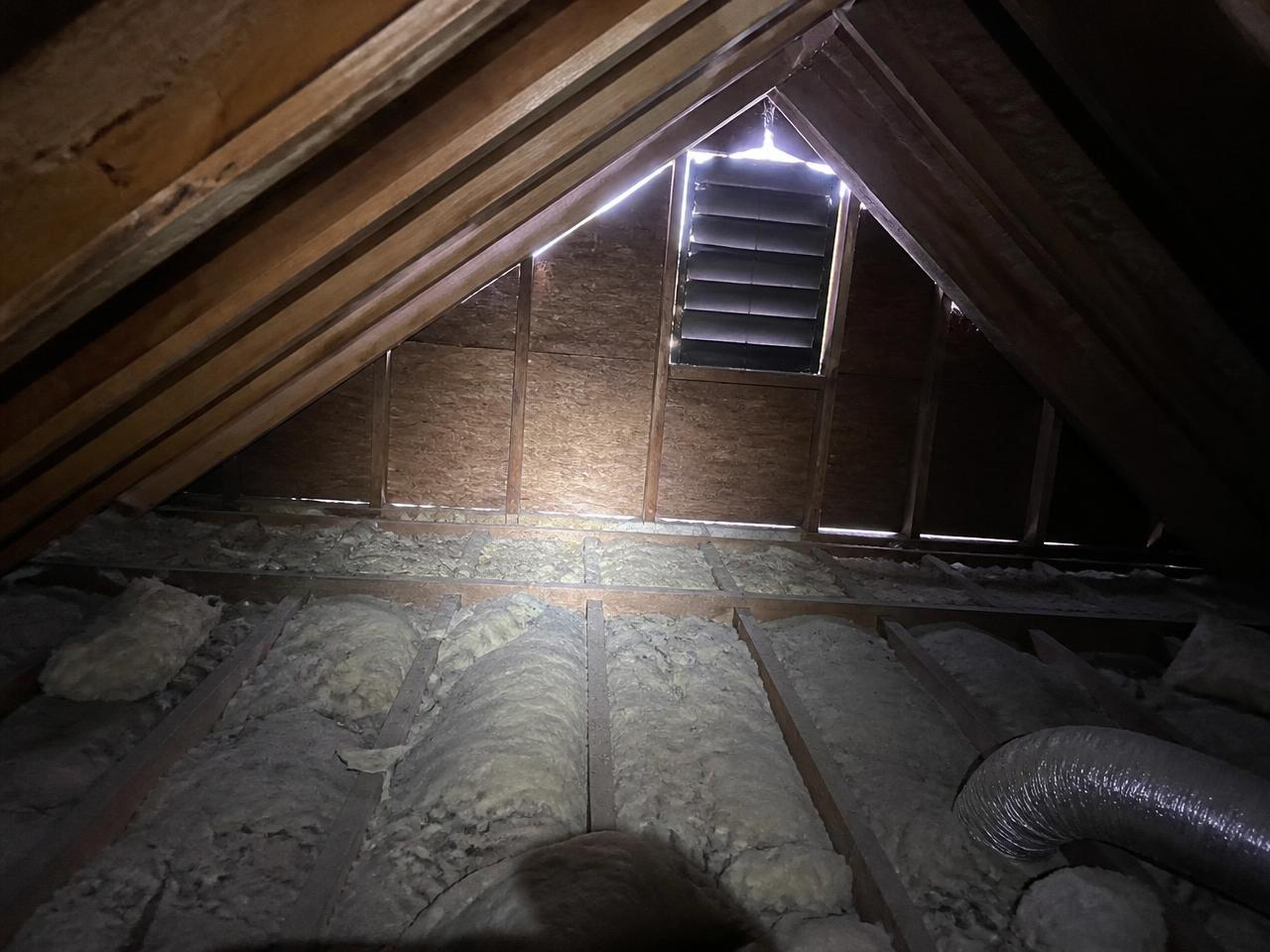 Flashlight-lit attic interior showing roof decking, rafters, a gable vent with daylight around its edges and old fiberglass insulation